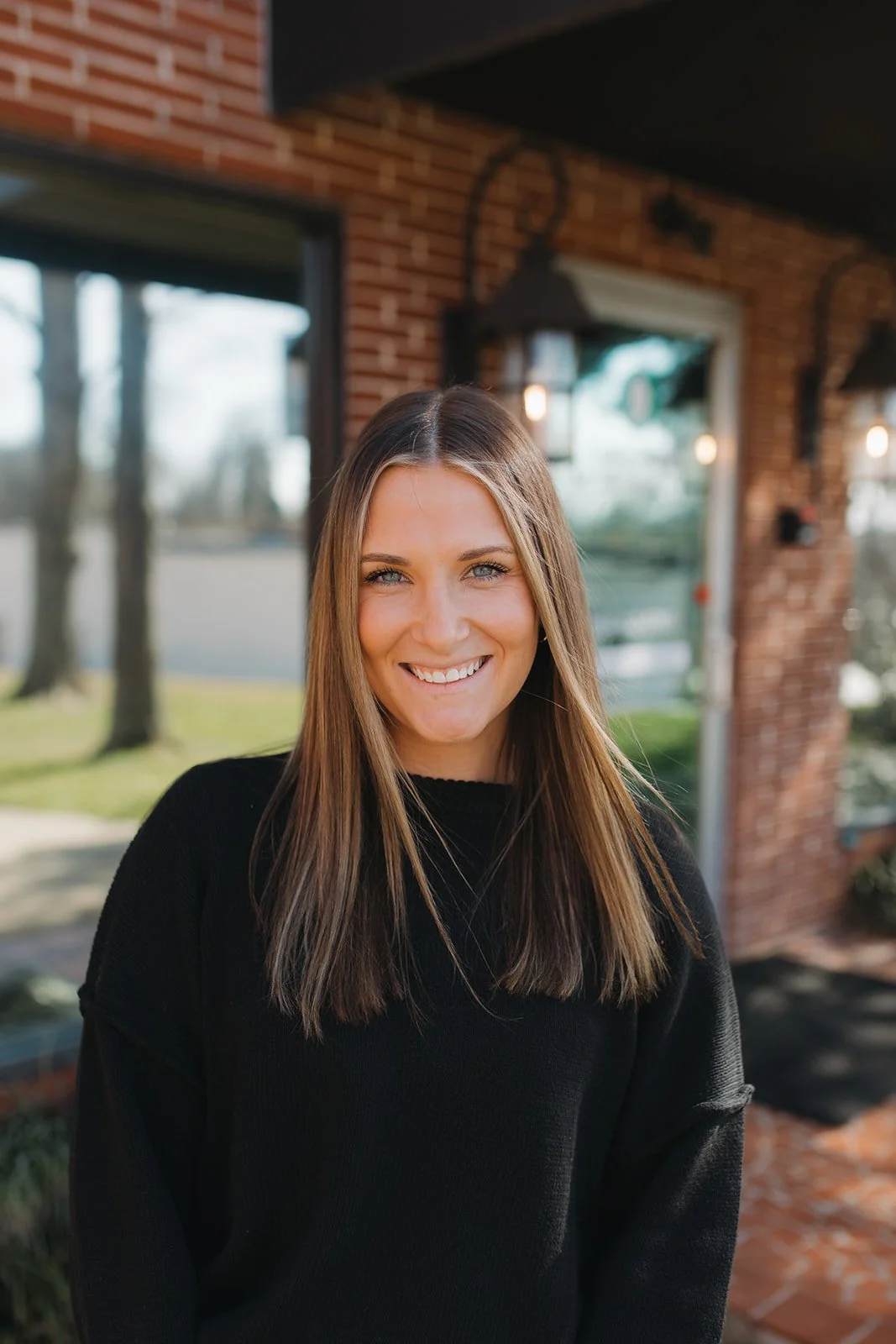 A woman with long brown hair smiling outdoors in front of a brick building, wearing a black sweater.