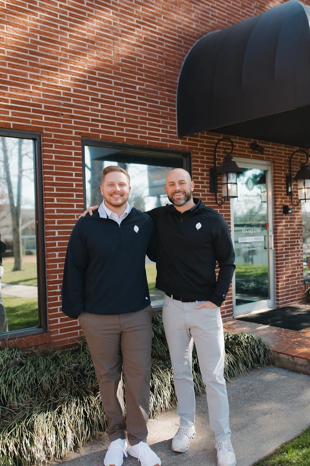 Two men standing side by side outside a brick building with large windows, smiling. One has his arm around the other's shoulder. Both are wearing black jackets with white leaf logos, beige pants, and white sneakers. The building has black awnings and outdoor wall lanterns.