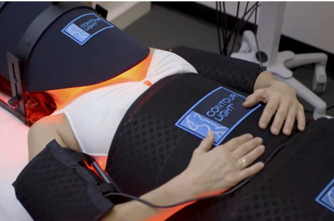 Person lying on a Contour Light therapy bed with orange light therapy pads, with hands resting on her abdomen, in a medical or therapy setting.