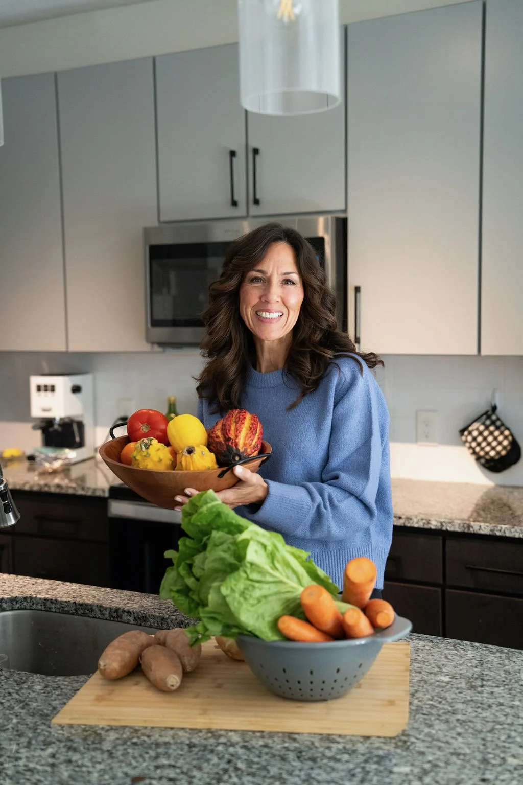 Woman in a blue sweater holding a bowl of colorful vegetables in a modern kitchen.