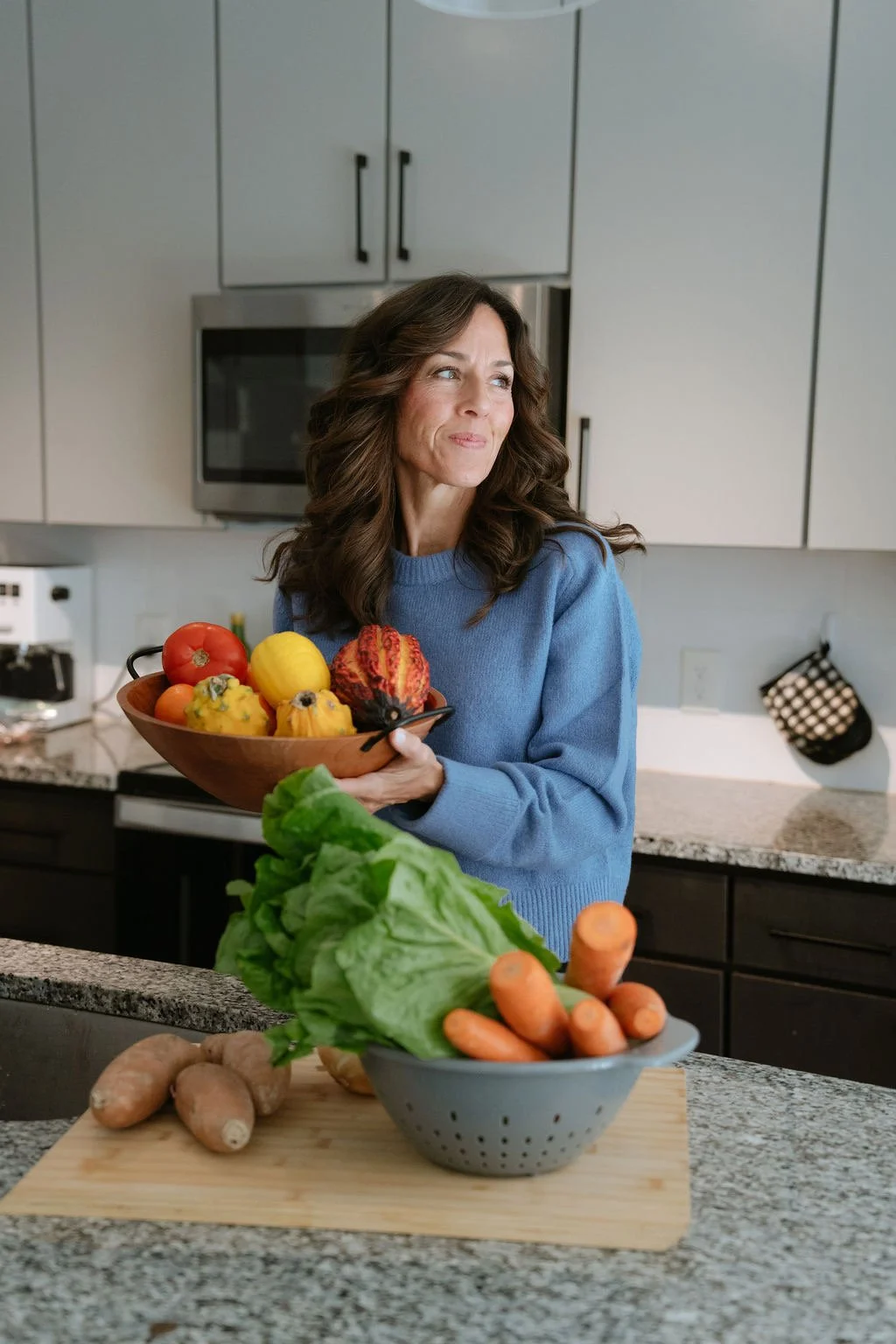 A woman holding a wooden bowl of colorful gourds and small pumpkins in a modern kitchen with gray cabinets and granite countertops.