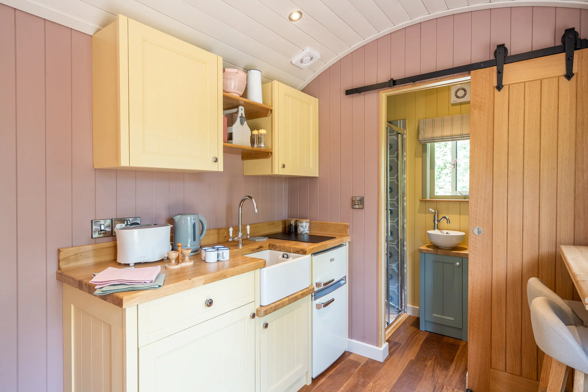 A cozy kitchen with pink and yellow walls, wooden accents, a small sink, a window with a view of outdoor greenery, a sliding barn door, and various kitchen appliances and utensils on the countertop.