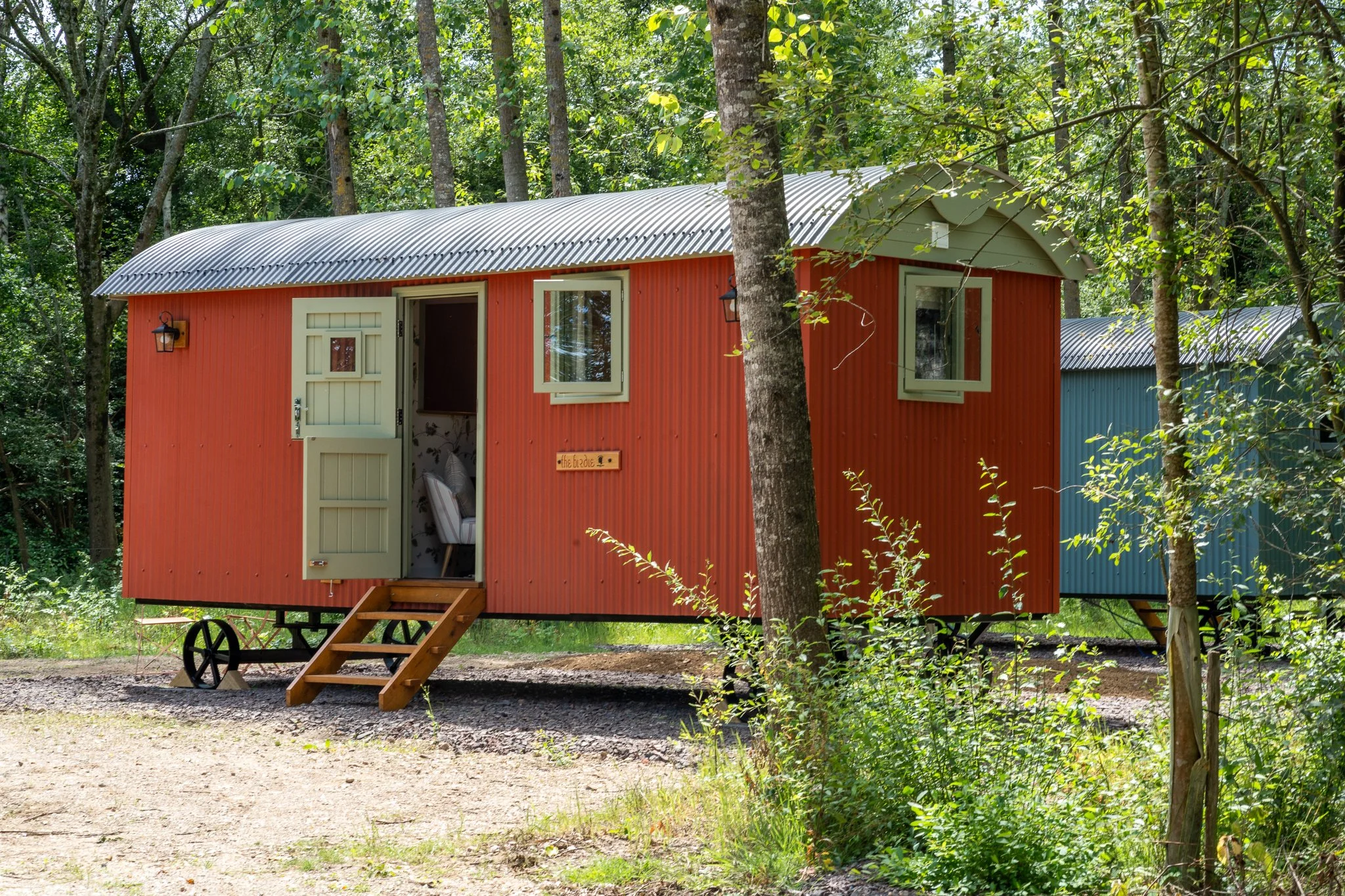 A small, red, B&B Shepherd Hut with a corrugated metal roof, set among green trees, with a small staircase leading to the entrance.