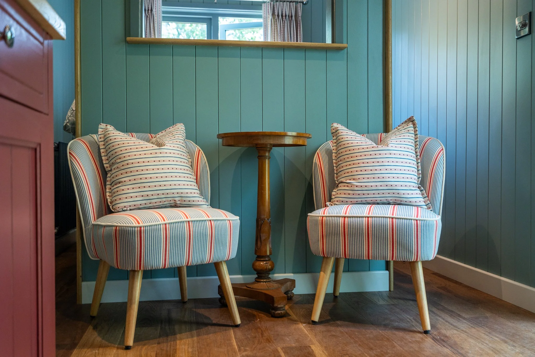 Two striped upholstered chairs set around a small wooden table in a cozy corner of a room with green paneled walls and a window with floral curtains.