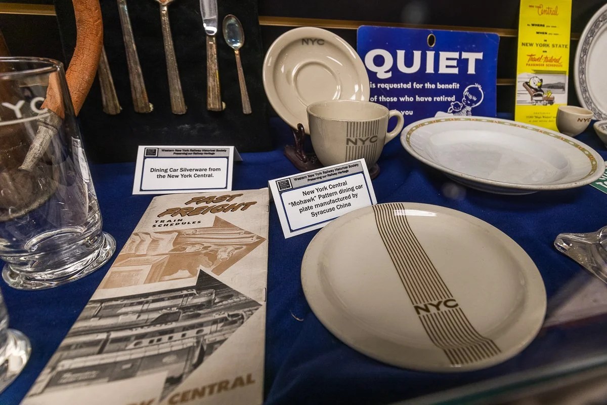 Display of vintage dining ware, including plates, cups, a glass, and informational signs about items from the New York Central railroad, on a blue cloth.