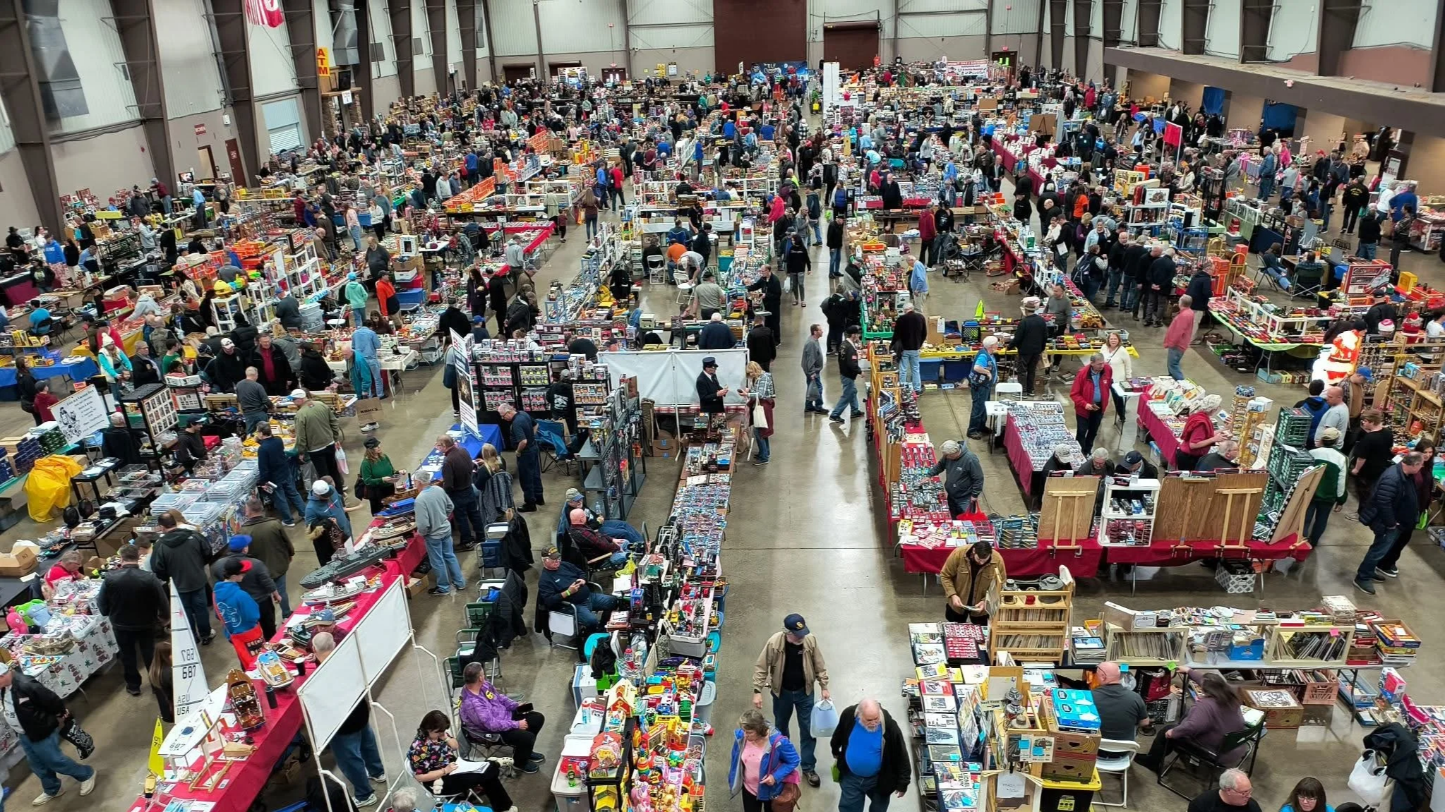 A crowded indoor flea market or swap meet with numerous tables filled with various items for sale, and many shoppers browsing.