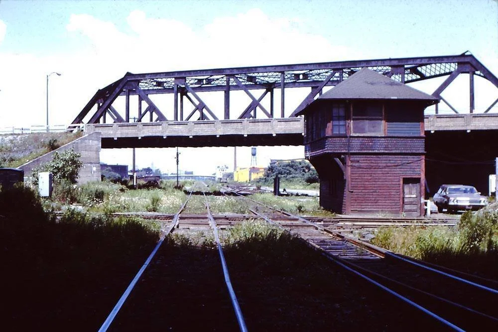 View of train tracks leading towards a bridge and a wooden control tower beside the bridge, with cars parked nearby.