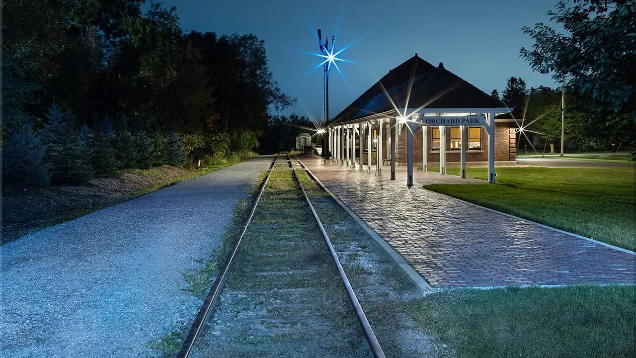 Night view of Orchard Park train station with lit platform, train tracks, and surrounding trees.