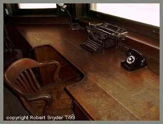 Wooden desk with an antique typewriter and a rotary phone, next to a wooden chair.