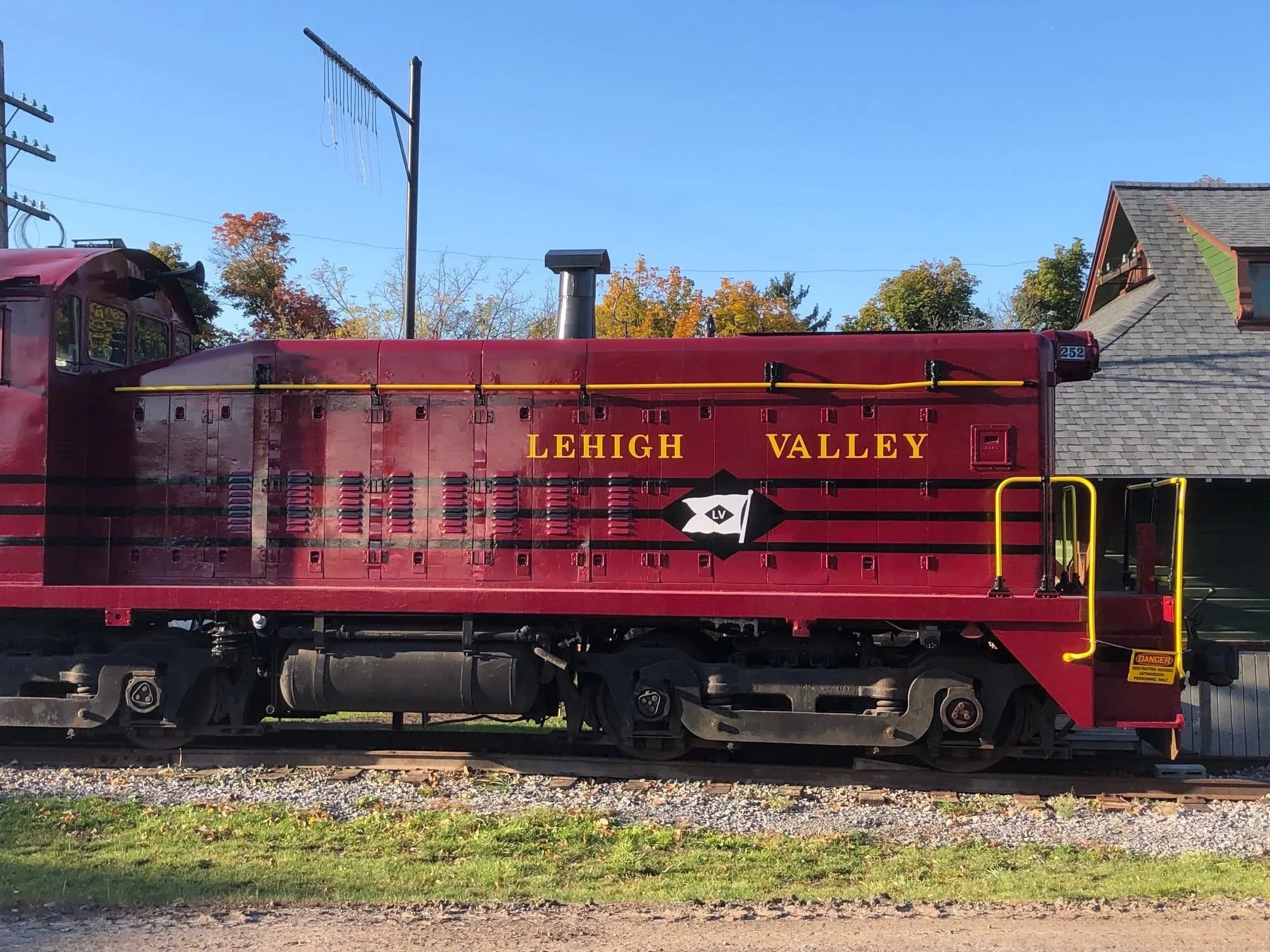 A red locomotive with the words 'LEHIGH VALLEY' written on its side, situated on railway tracks with autumn-colored trees and houses with pitched roofs in the background under a clear blue sky.