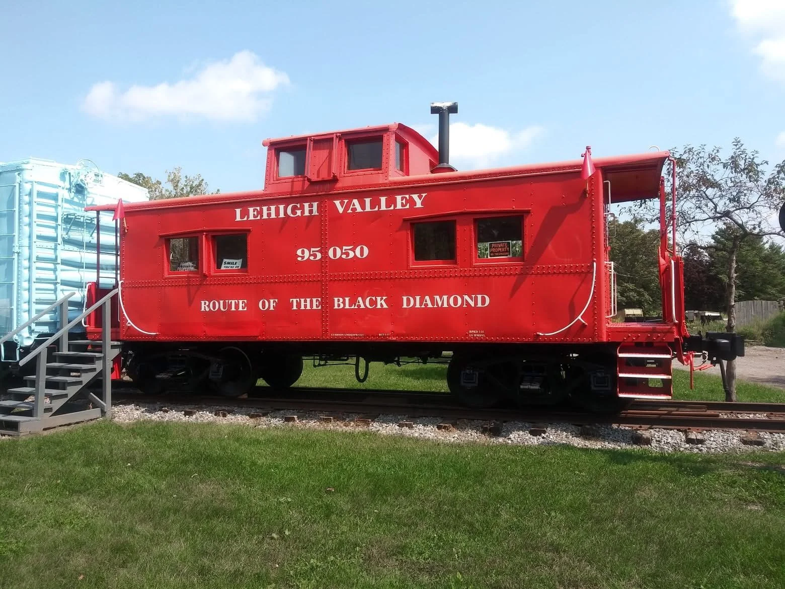 Red historic train car with white lettering reading 'Lehigh Valley Route of the Black Diamond,' positioned on railway tracks, with a small set of metal stairs leading to it and trees in the background under a partly cloudy sky.