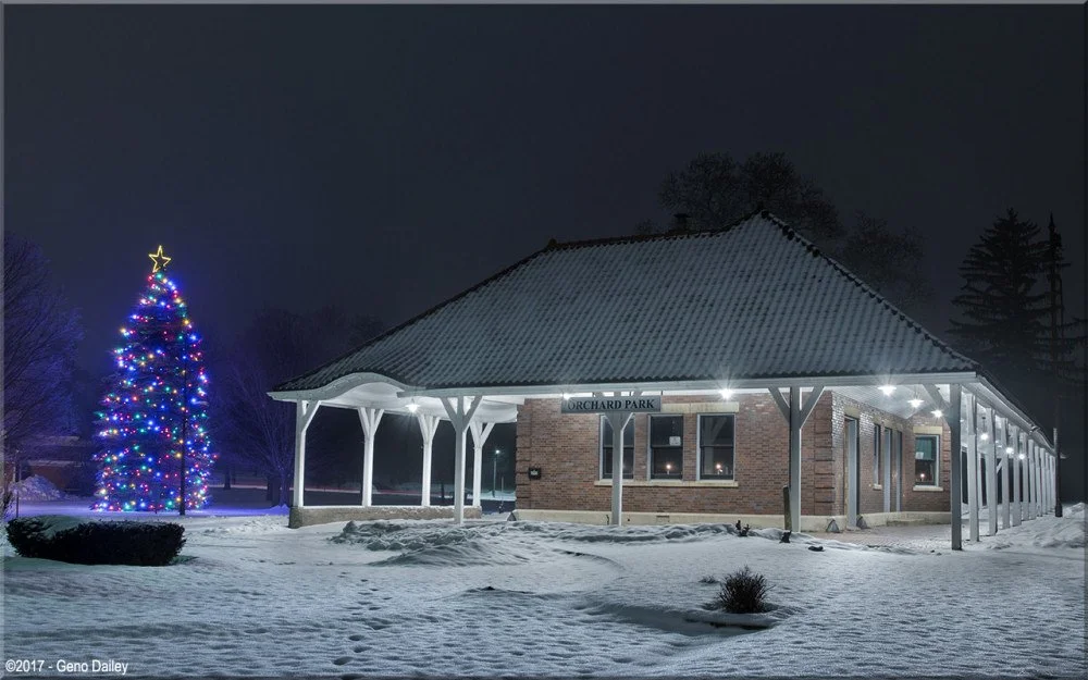 Snow-covered Orchard Park train station at night with a decorated Christmas tree lit with colorful lights and a star on top.