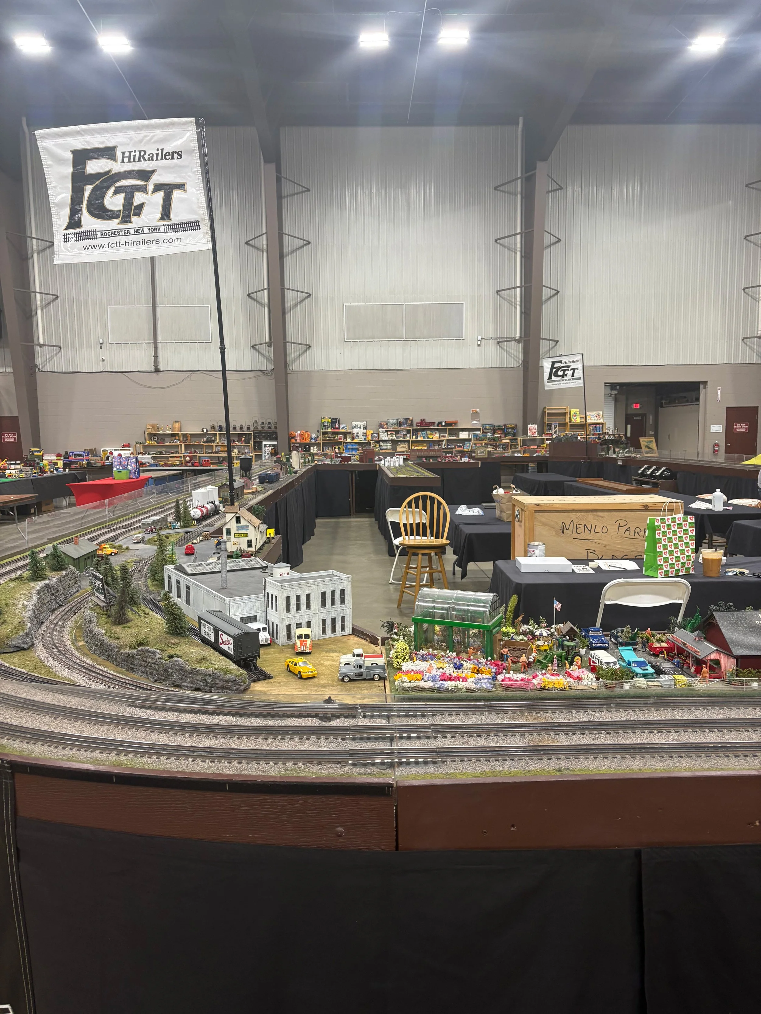 Model train display with miniature buildings, vehicles, trees, and tracks in an indoor exhibition space.