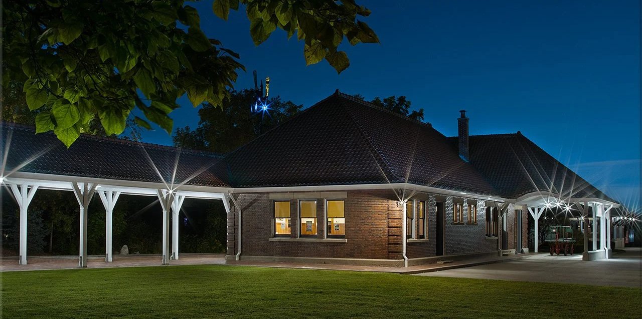 Night view of a brick building with windows, surrounded by a white pergola structure with lights, and topped with dark roof tiles.