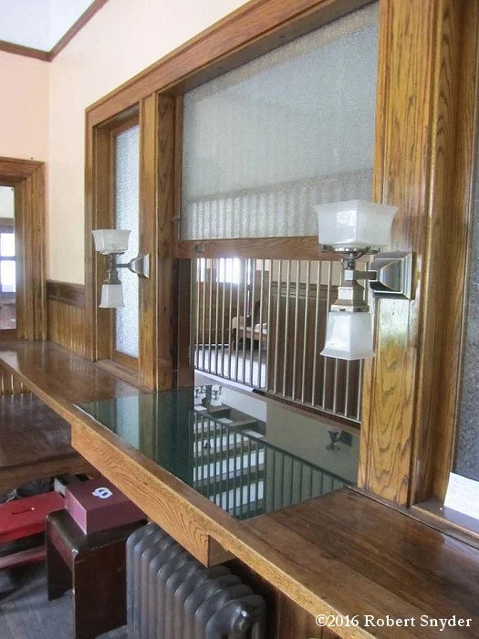 A wooden ledge with two wall-mounted lamps, a large window with a frosted glass upper pane, and a mirror in the middle reflecting a room with a stair railing.