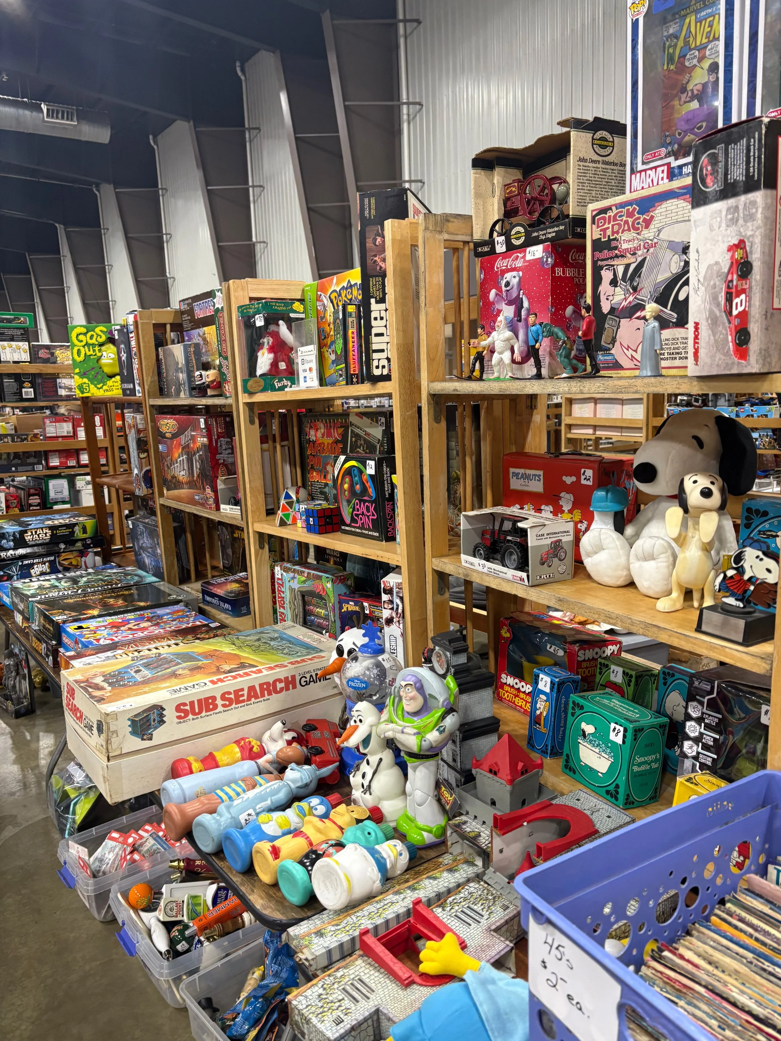 A wide view of a thrift store shelf display with various toys, games, and collectibles including plush toys, action figures, board games, and comic books.