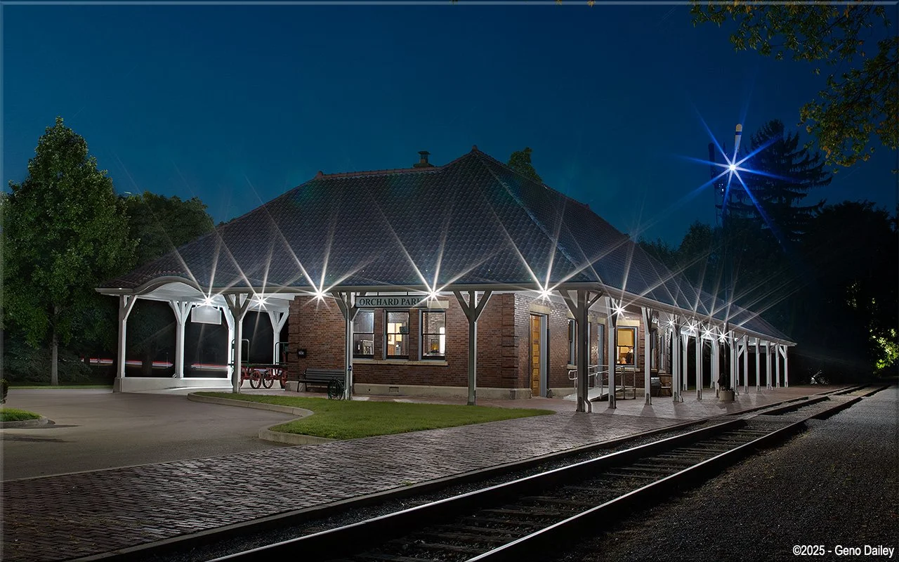 Nighttime view of a train station with a brick building, illuminated by bright exterior lights, with train tracks in the foreground and trees in the background.