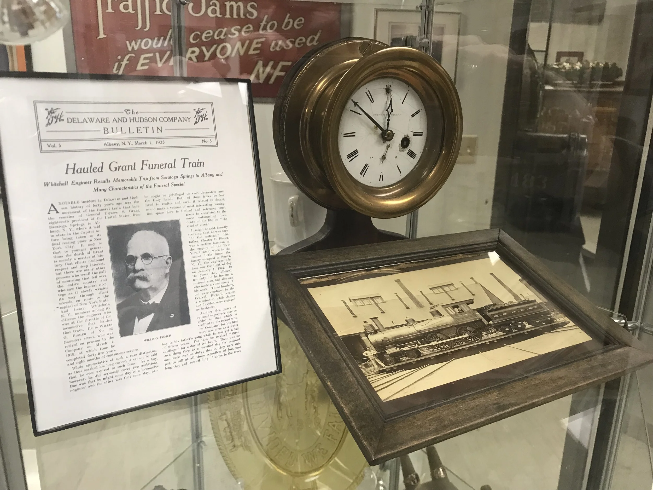 An antique clock with a brass frame displaying Roman numerals, positioned inside a glass case. Beside it, there is a framed black-and-white newspaper article about the Delaware and Hudson Company, featuring a photo of a man in a tuxedo with a bow tie