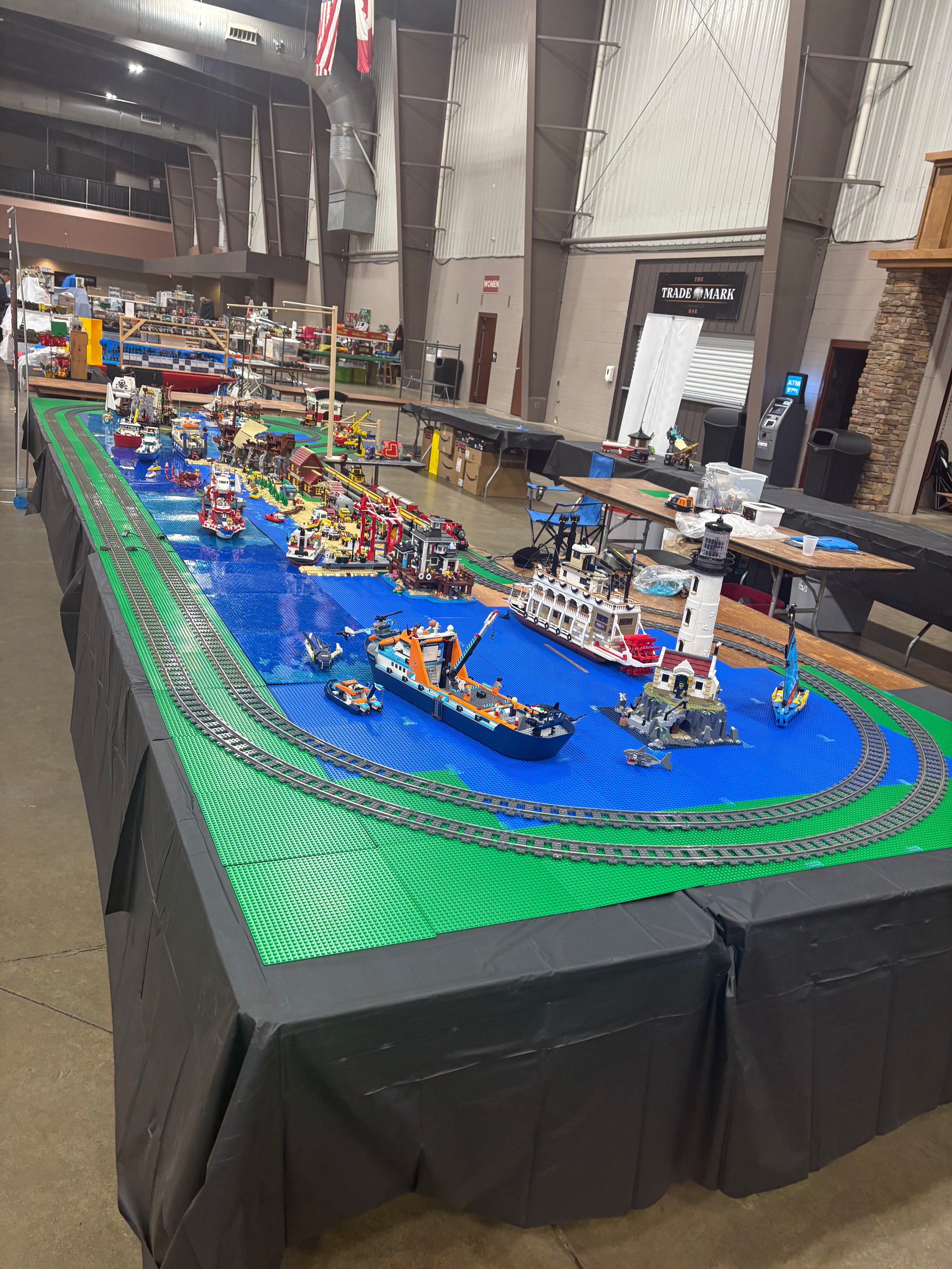 Table with a detailed LEGO cityscape, featuring boats, buildings, and a train on a track, set up indoors with a black tablecloth.