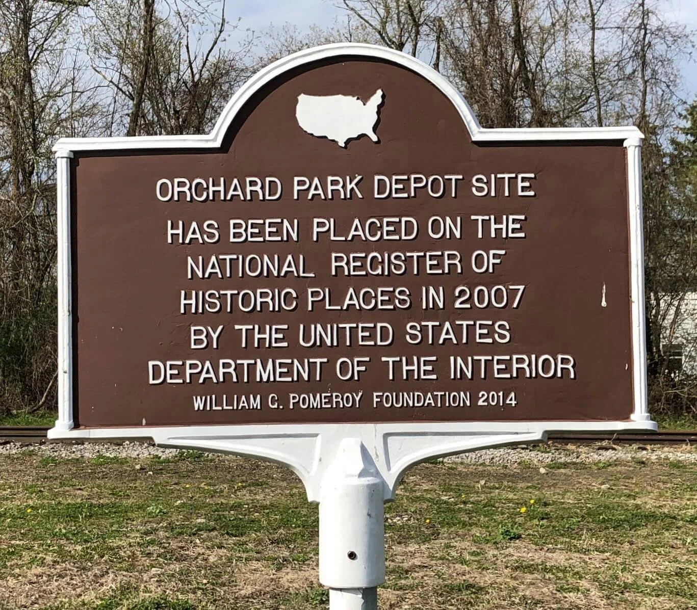 Historic brown and white signboard for Orchard Park Depot Site, with an outline of the United States at the top, indicating its addition to the National Register of Historic Places in 2007 by the U.S. Department of the Interior, funded by William G. Pomeroy Foundation in 2014, situated outdoors with trees in the background.