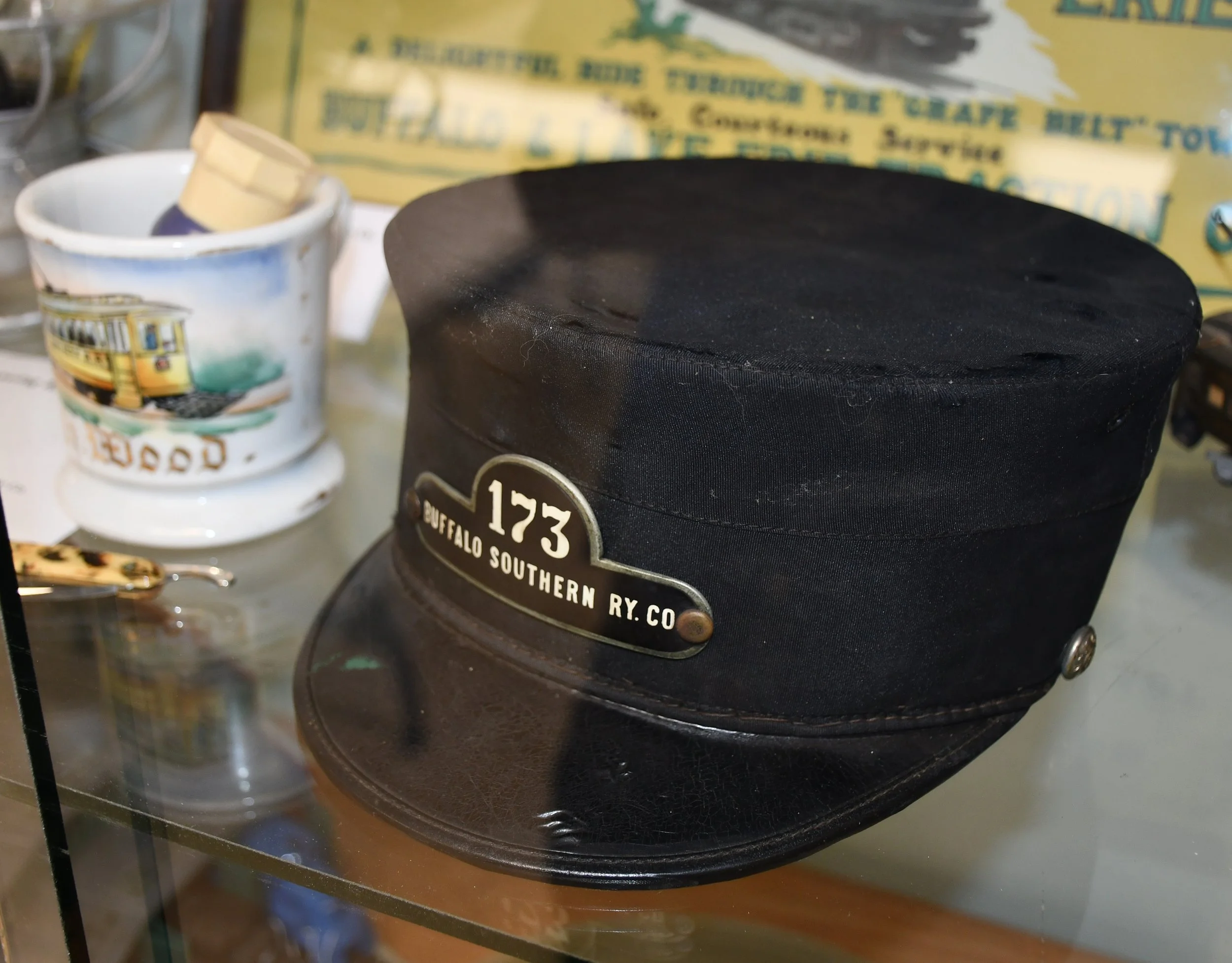 Close-up of a black hat with a tag that reads "123 Buffalo Southern Ry Co." on a reflective surface, with a mug featuring a train illustration and a yellow box in the background.