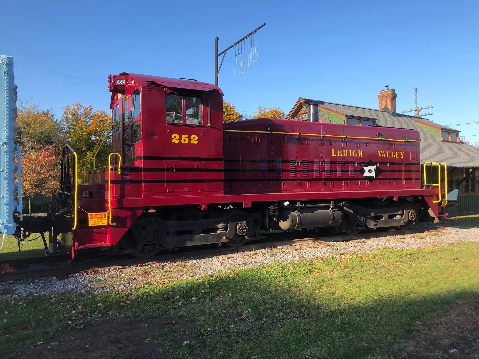 A vintage red train engine with the words 'Lehigh Valley' and the number 252, parked on train tracks in a suburban area with fall trees and a house in the background.