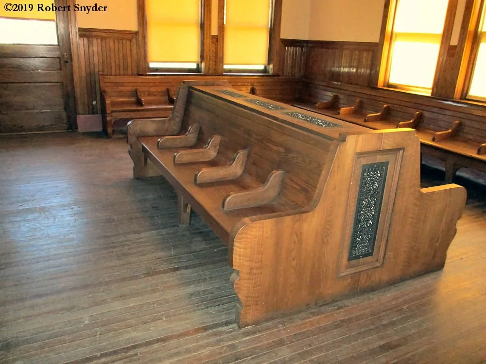 An empty wooden room with a church pew and a confessional booth in the center. The room has wooden walls, floors, and windows with yellow curtains.
