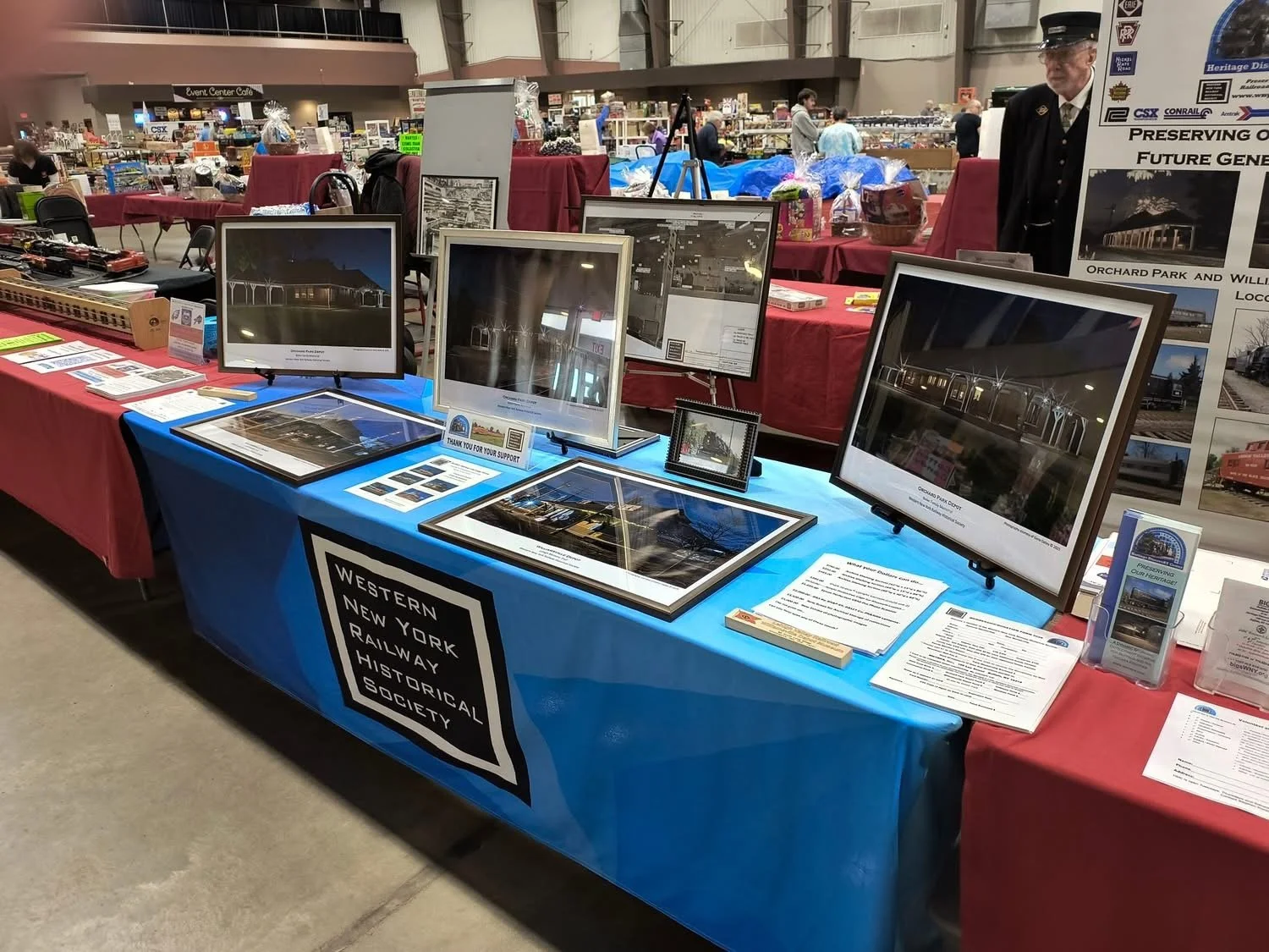 Exhibit table with framed photos and informational materials representing the Western New York Railway Historical Society at an indoor event, with other tables and attendees in the background.