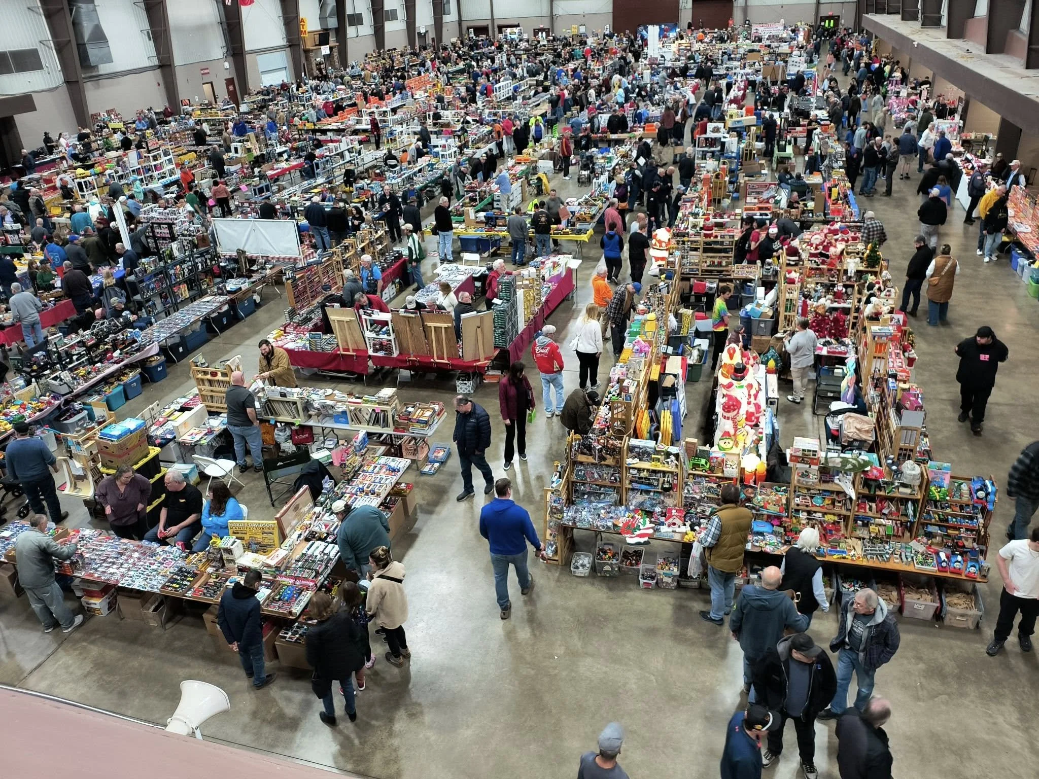 A crowded indoor flea market or swap meet with many tables filled with various items for sale, including toys, books, and collectibles. People browse and shop among the multitude of vendors.