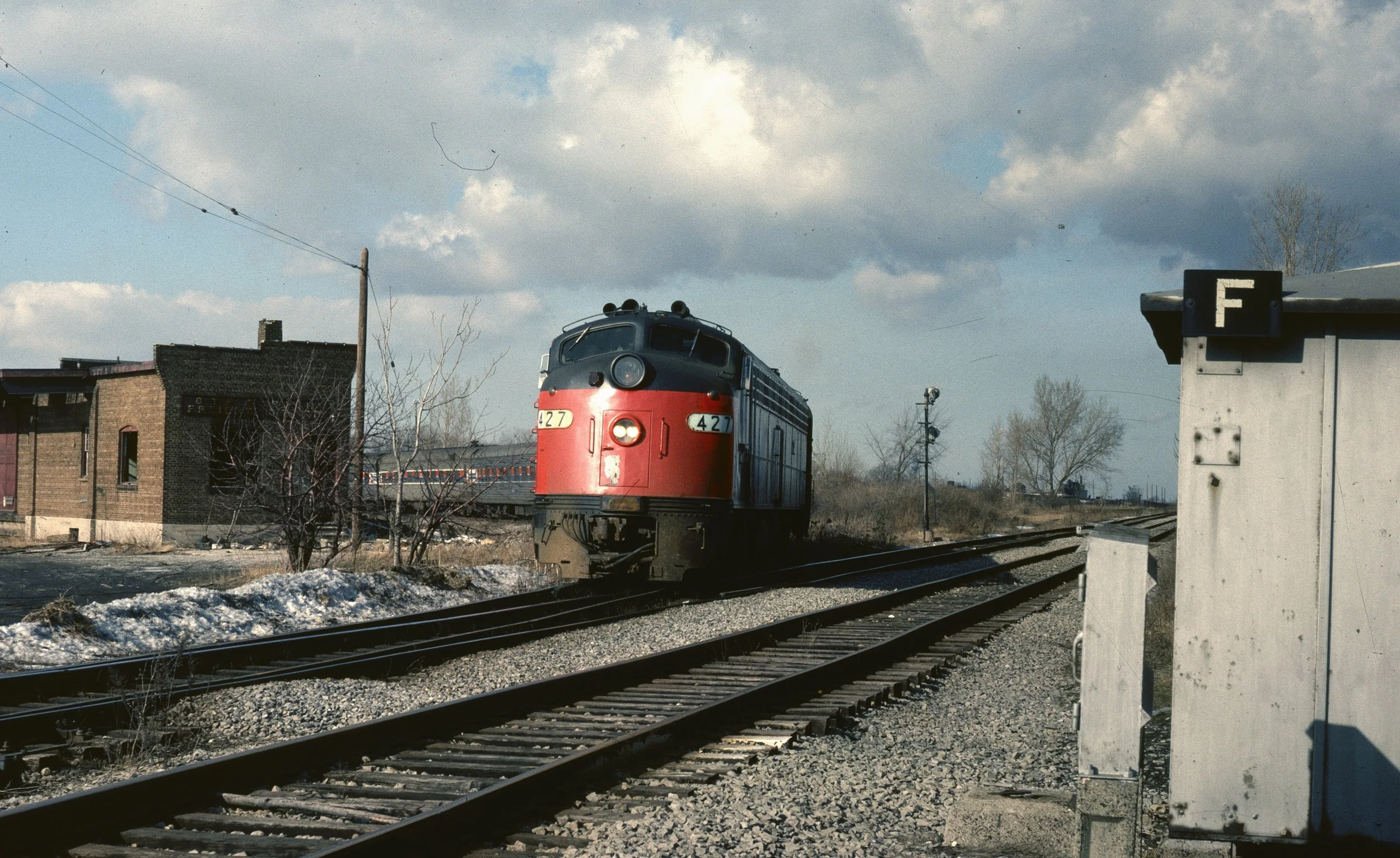 A red and black diesel train traveling on a railway track in a rural area with some leafless trees, buildings, and utility poles visible. The sky is partly cloudy.