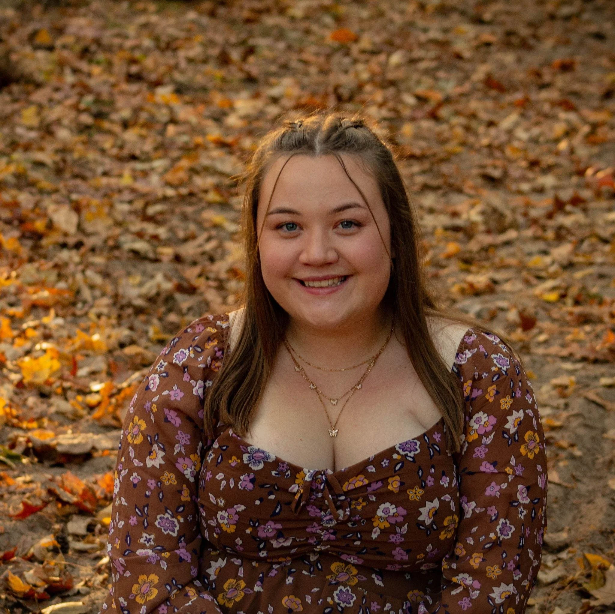 A young woman with brown hair wearing a floral brown dress, smiling, in an outdoor setting with autumn leaves.