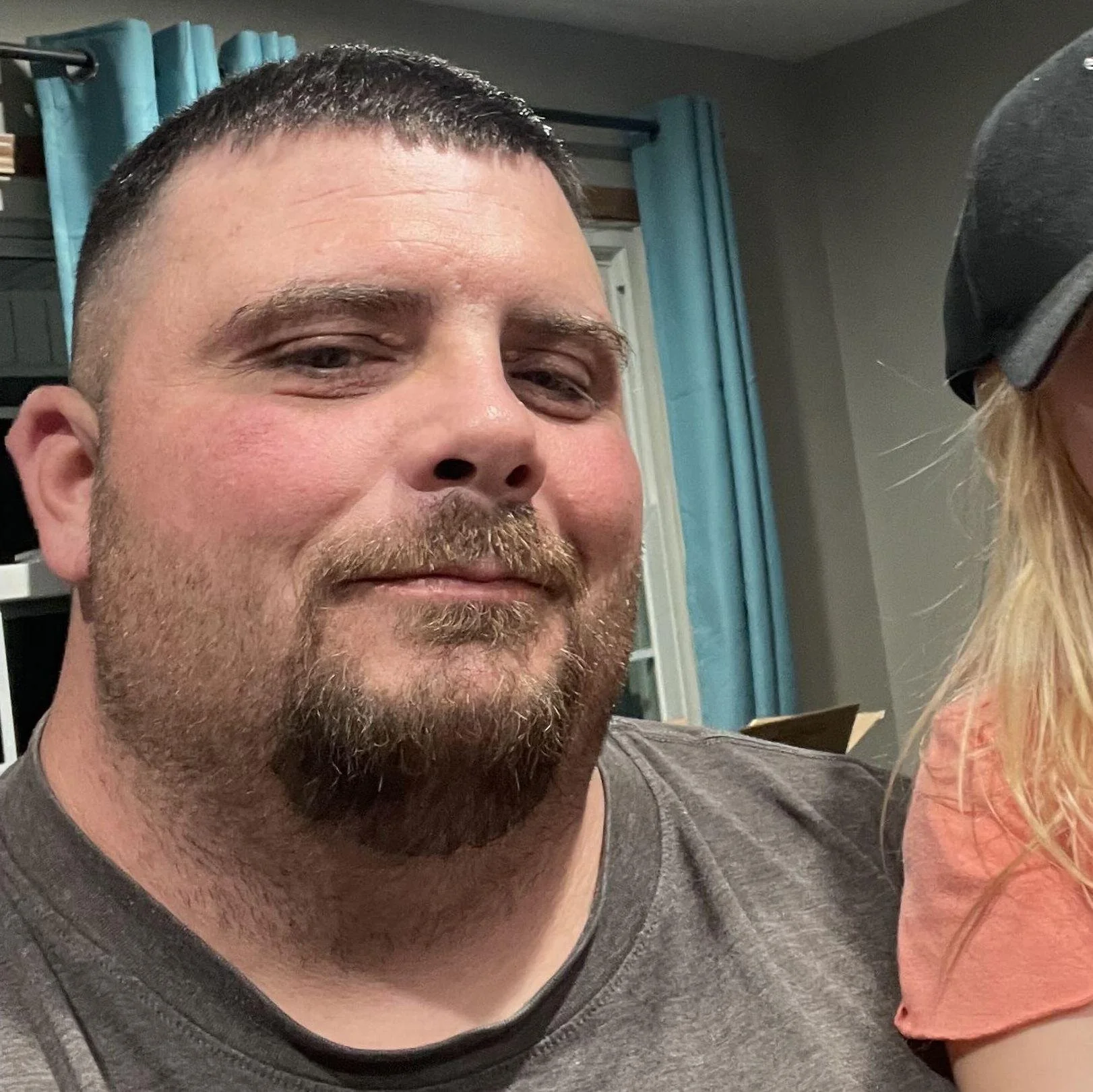 Close-up of a man with short dark hair, a beard, and a mustache, wearing a gray T-shirt, in a room with blue curtains and a window in the background.