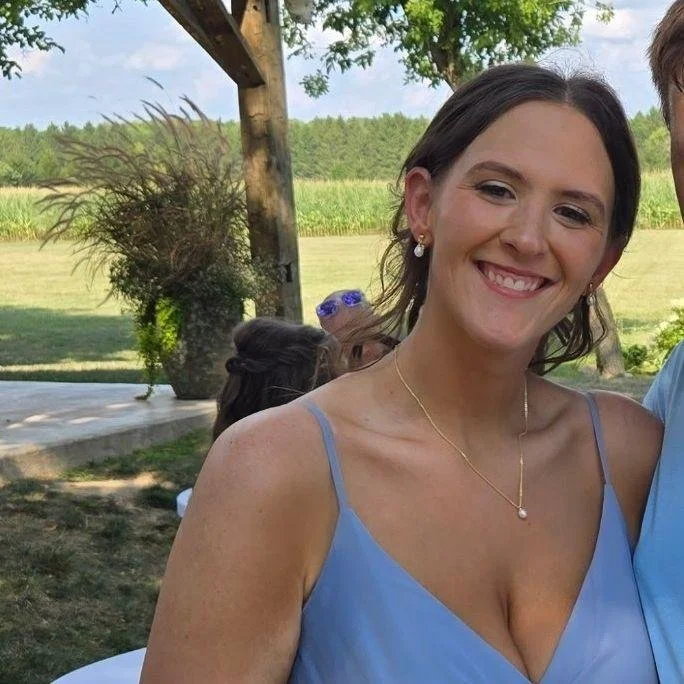 A smiling woman with dark hair in a bun, wearing a light blue dress with spaghetti straps, standing outdoors near a tree and greenery.