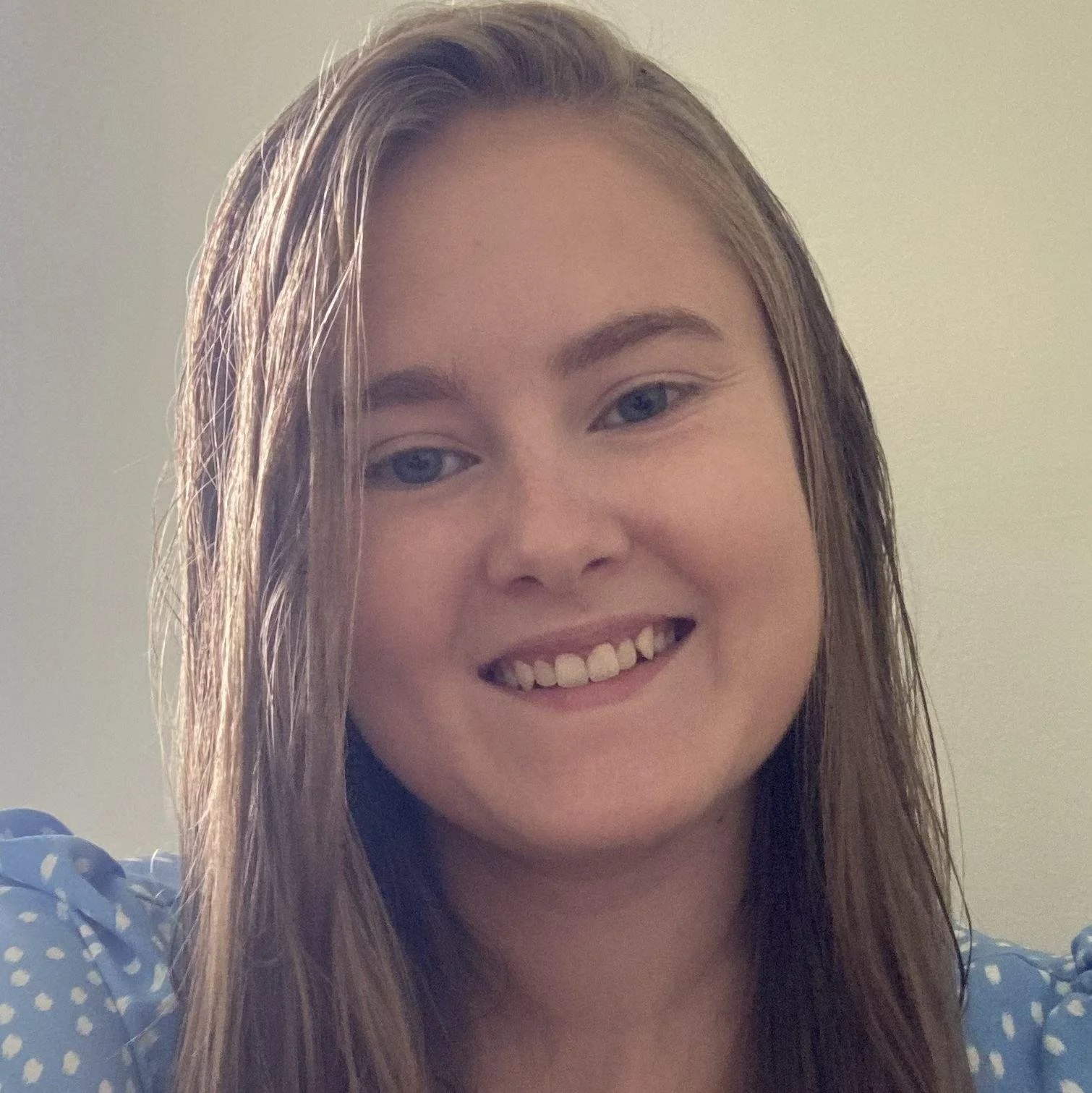 Close-up photo of a young woman with wet, light brown hair, smiling, wearing a blue polka dot top, against a plain light-colored background.