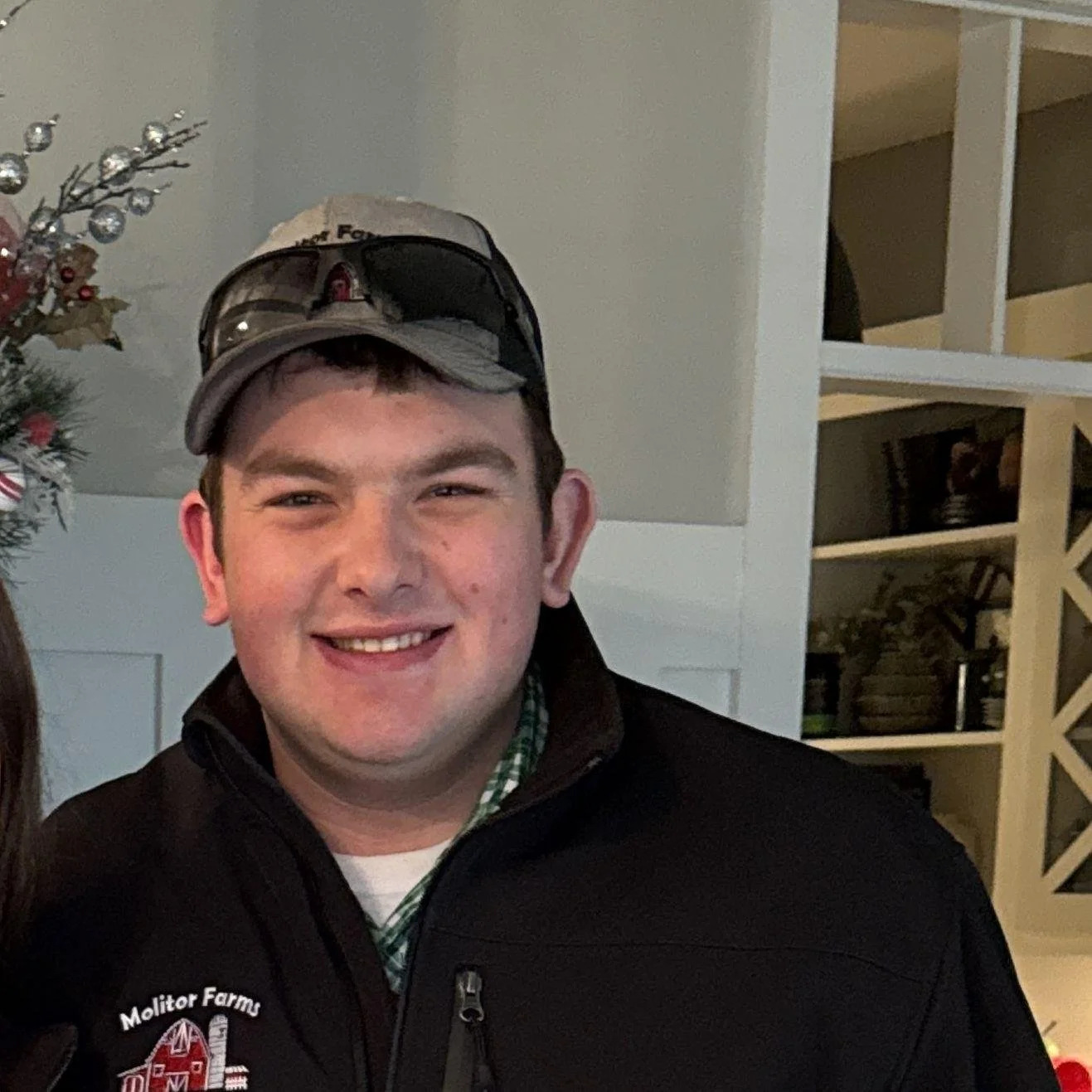 A young man smiling, wearing a baseball cap and a black zip-up jacket with an embroidery logo of Molitor Farms. There is a Christmas tree with ornaments on the left side of the image. The background includes a white shelf and wall with room decor.