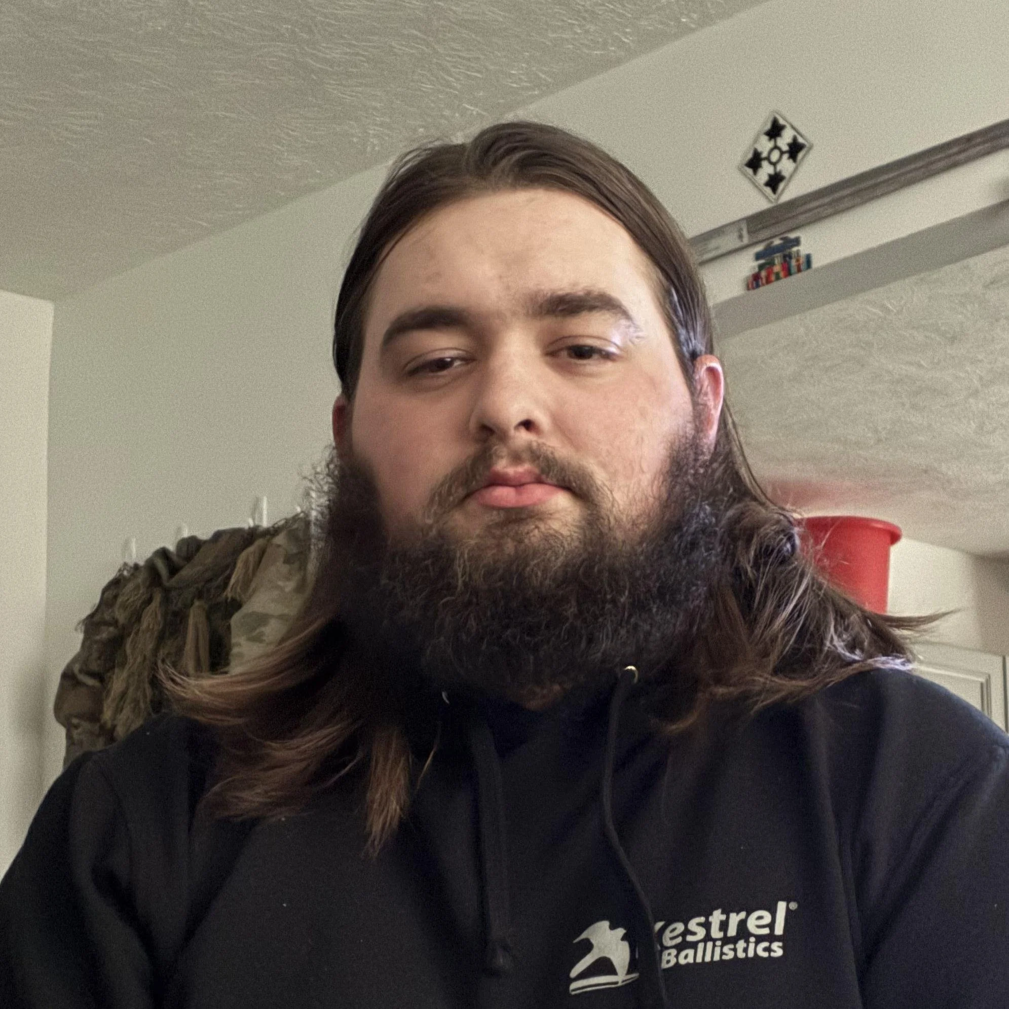 A young man with long hair and a beard taking a selfie indoors, wearing a black Westrel Ballistics hoodie. The background shows a ceiling, a shelf with small items, and a red container.