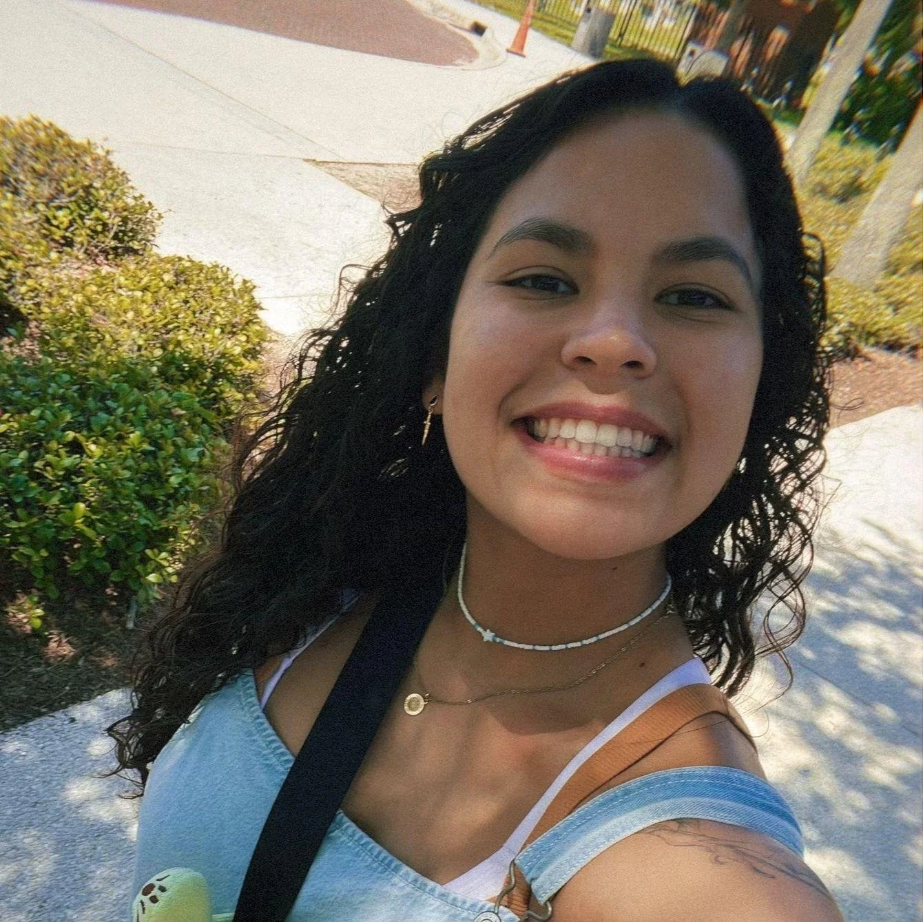 A woman smiling outdoors with trees and a sidewalk in the background.