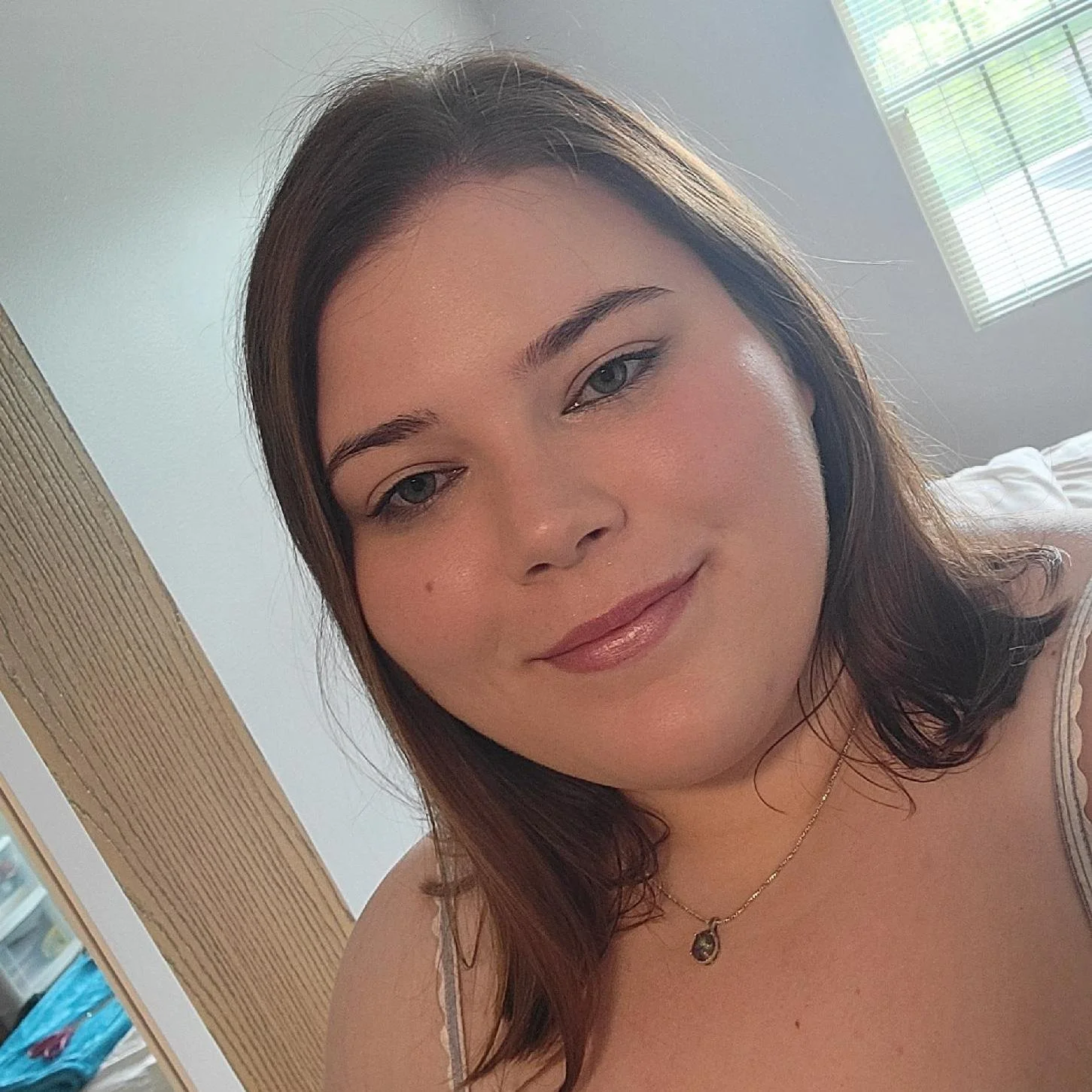 A woman with shoulder-length brown hair, light skin, and blue eyes smiling softly. She is wearing a gold necklace with a pendant and a striped tank top. The background shows a light-colored wall, a window with blinds, and part of a bed.