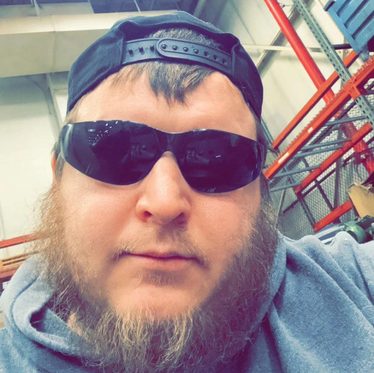 A man with a beard wearing black sunglasses, a backwards black cap, and a gray hoodie, in an industrial or warehouse setting with orange shelves and metal racks in the background.