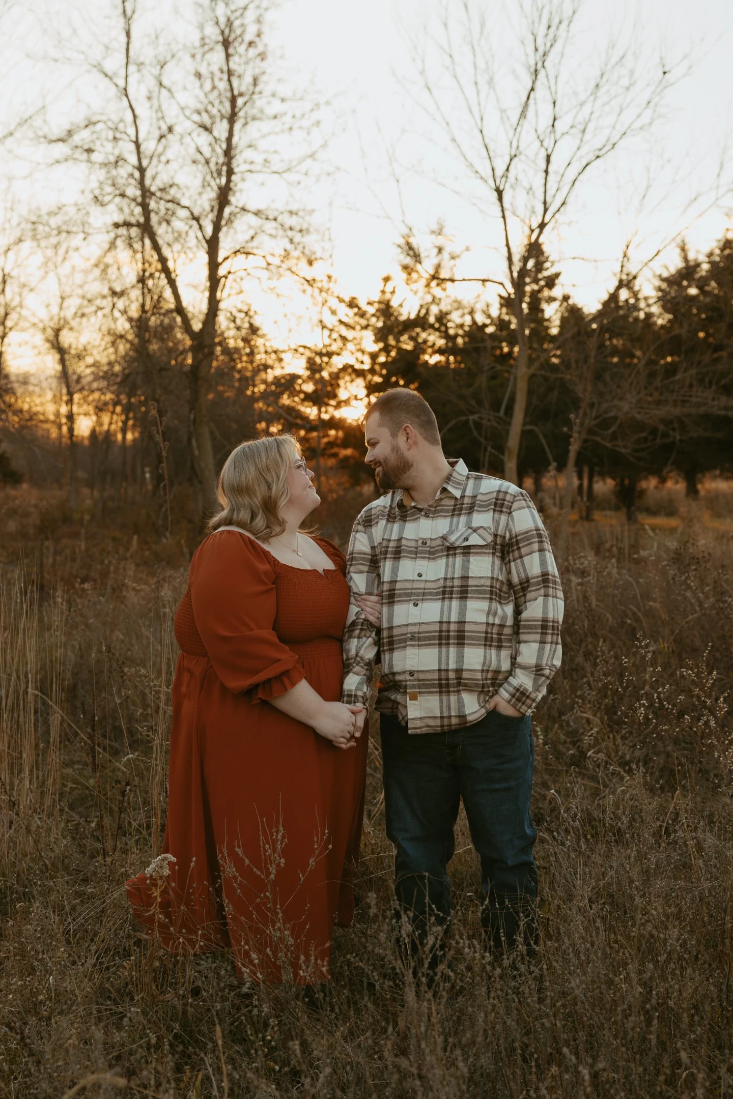 A couple standing in a field during sunset, holding hands and gazing at each other, with bare trees in the background.