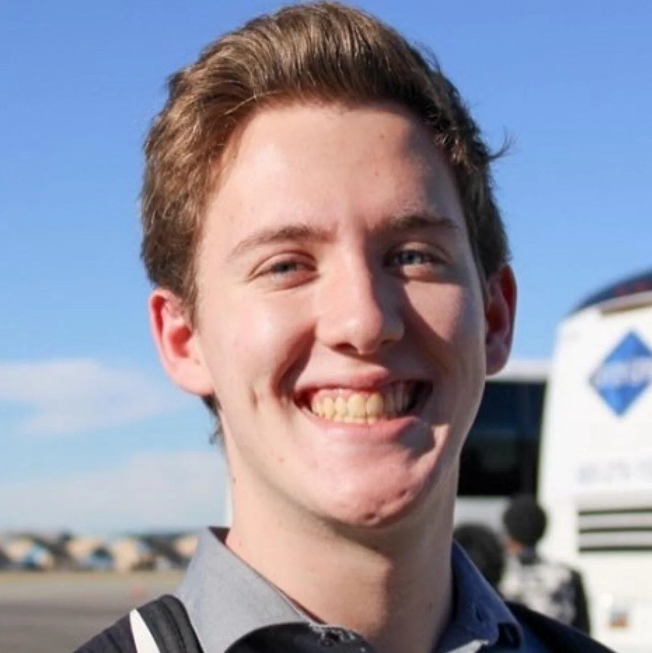 Close-up of a young man smiling outdoors with a clear blue sky and a bus in the background.