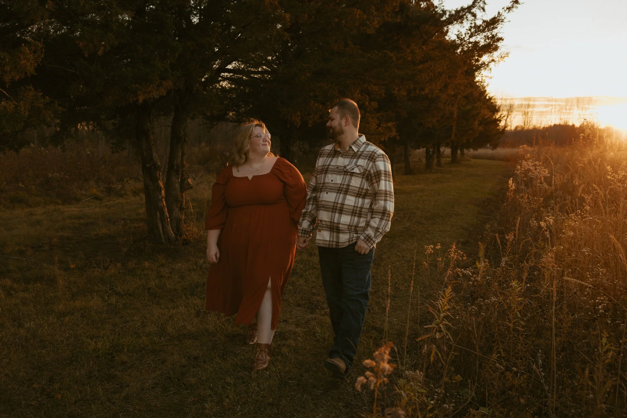 A couple walking hand in hand on a grassy trail at sunset, surrounded by trees and wildflowers.