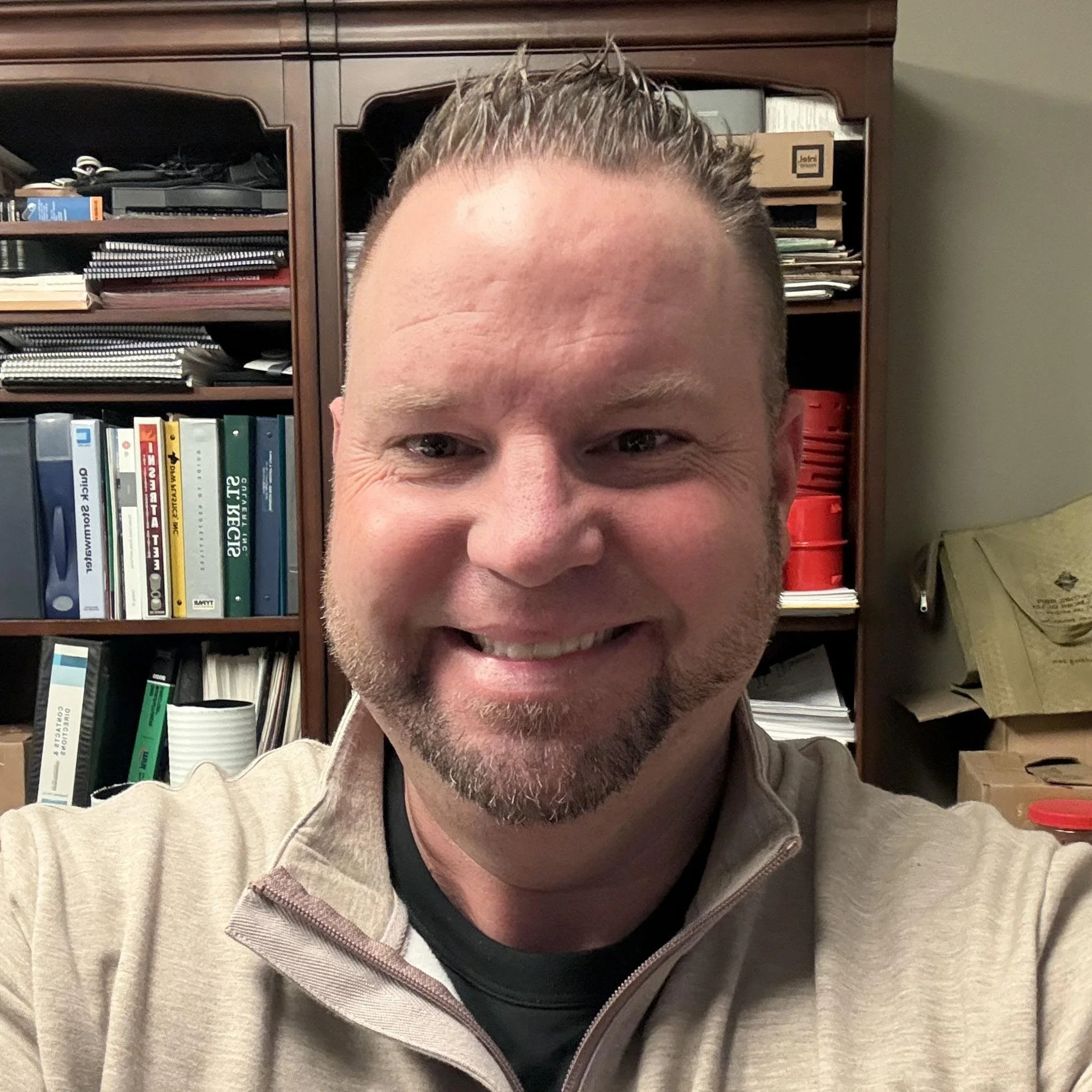 A smiling man with short hair and facial hair taking a selfie in an office filled with books and office supplies.