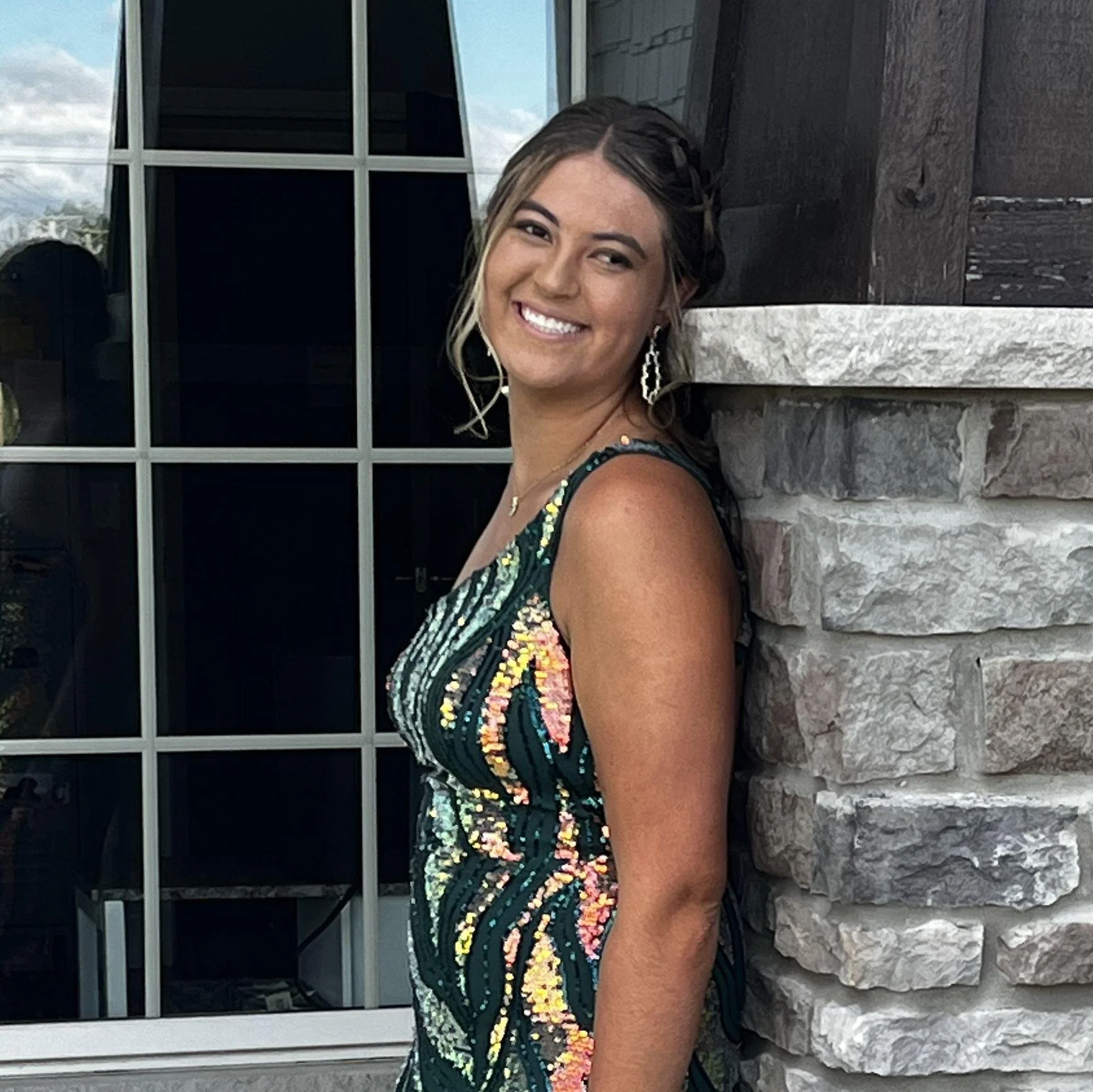 Smiling young woman with long hair in an elegant dress standing against a stone wall outside a building with large windows.
