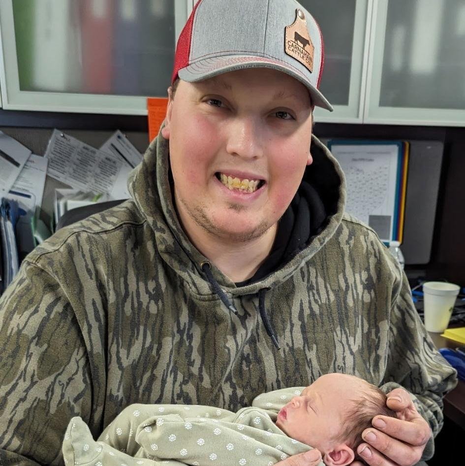 A young man wearing a gray and red cap and a camouflage hoodie is holding a newborn baby wrapped in a light green blanket with white dots. They are in an office environment with papers, a monitor, and a cup in the background.