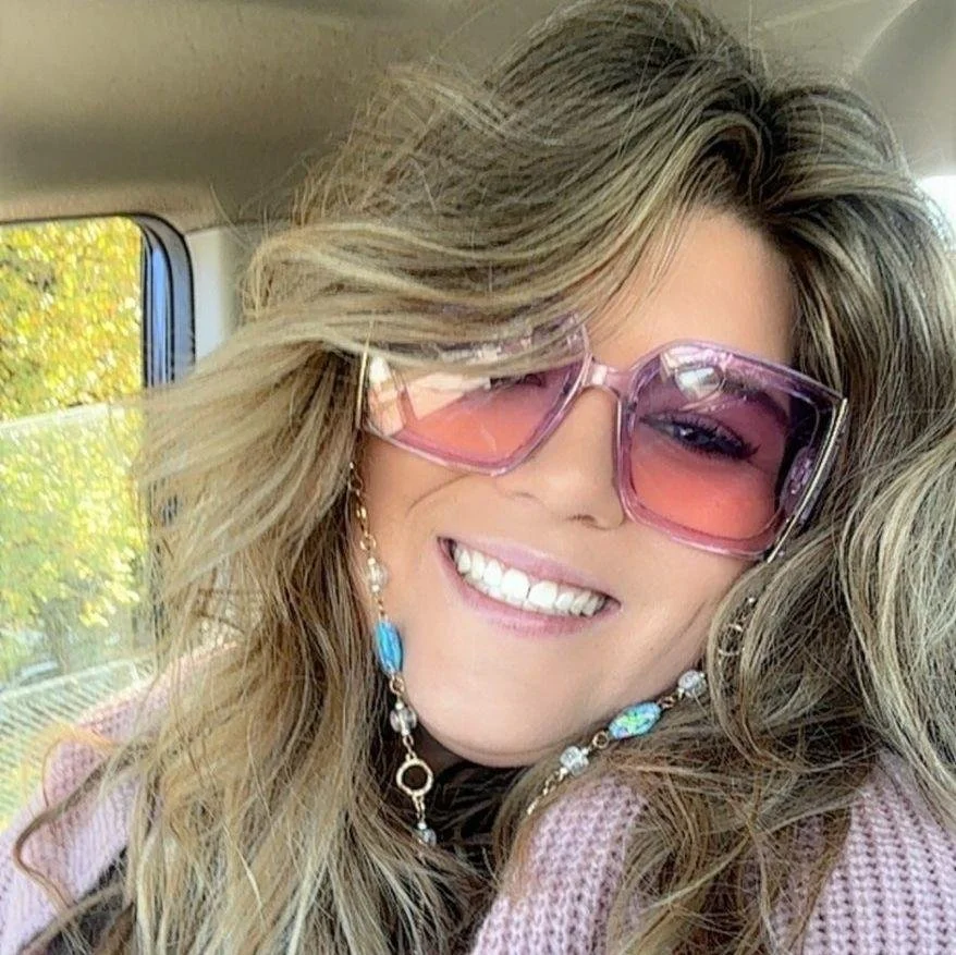 A woman with long wavy blonde hair wearing pink sunglasses, smiling showing her teeth, and wearing colorful jewelry with a pink knit sweater. She is inside a vehicle with a window showing trees outside.
