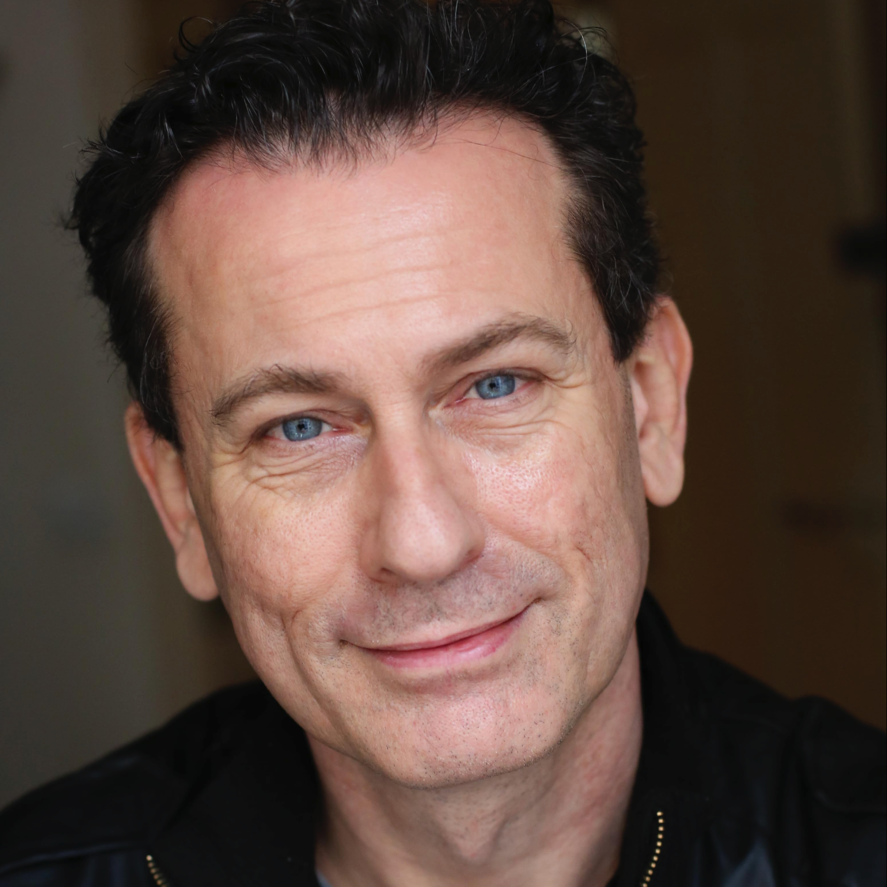 Close-up of a smiling middle-aged man with blue eyes, dark curly hair, and a black top, against a blurred background.