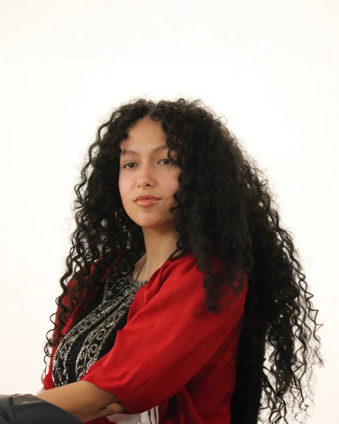 A woman with long, curly black hair wearing a red garment, sitting against a plain white background.