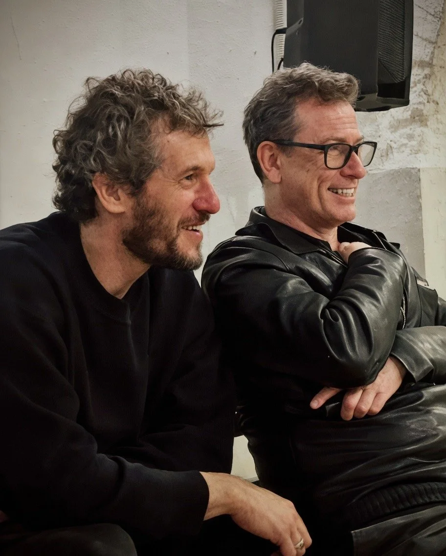 Two men sitting indoors, smiling and laughing, one with curly hair and beard, wearing a black shirt, and the other with glasses and gray hair, wearing a black leather jacket.