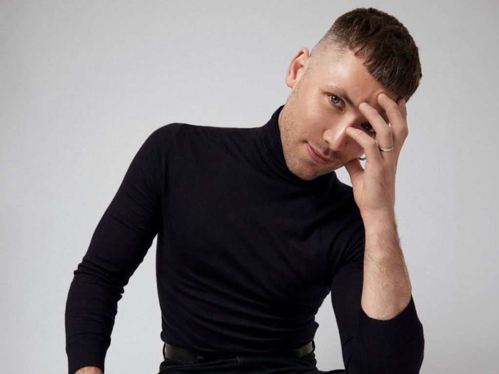 Close-up portrait of a smiling man with short dark hair, blue eyes, wearing a black turtleneck and a small hoop earring in his left ear, against a blurred neutral background.